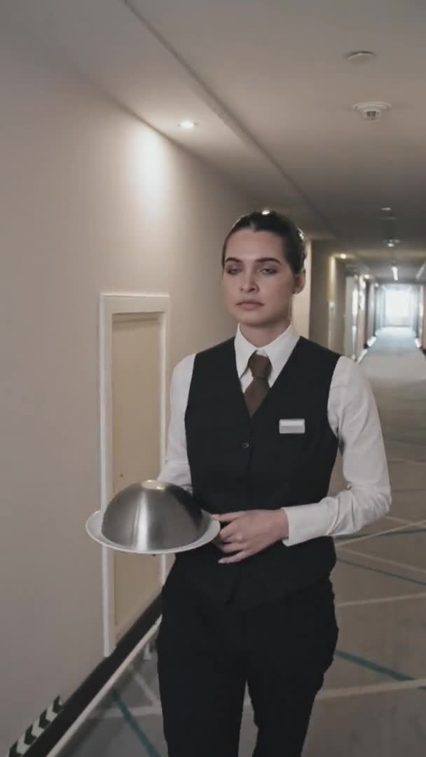 Vertikální záběr atraktivní mladé servírky kráčející po hotelové chodbě servírující jídlo na podnose s kovovou kupolí k otevíracím dveřím pro hosty