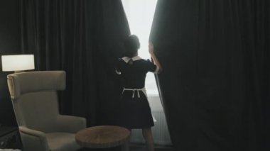 Modern otel odasındaki siyah perdeleri kapatan genç kadın hizmetçinin dikiz görüntüsü.