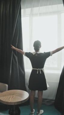 Dikey zoom-out, modern otel odasındaki siyah perdeleri açan siyah beyaz üniformalı kadın hizmetçinin yavaş çekimi.