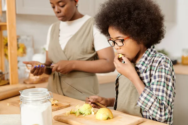 Netter Junge isst rohe Äpfel, während er Zutaten schneidet — Stockfoto