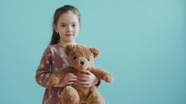 Küçük sevimli bir kızın portre resmi peluş ayı oyuncağıyla mavi arka planda dikilirken kameraya gülümsüyor.