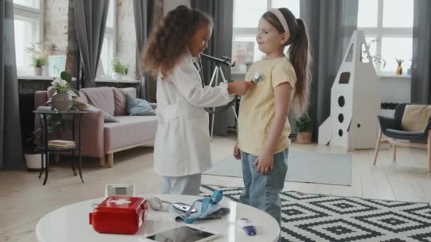 彼女の小さな友人を確認するために聴診器を使用して医師の衣装でかわいい8歳の女の子の手持ちの低速の肖像画 カメラに向かって笑っている — ストック動画