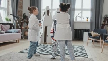 Laboratuvar önlüklü 8 yaşındaki kızların oturma odasında rol yaparken uzay giysisi giydiği orta boy bir fotoğraf. Arka planda duran mukavva roket gemisi
