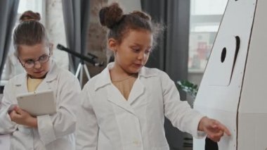 Evde oynarken karton roket gemisini inceliyormuş gibi yapan laboratuvar önlüklü iki küçük kızın el kamerasıyla çekilmiş yavaş çekimleri.