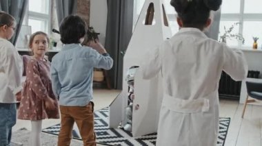 Heyecanlı küçük çocukların uzay elbiseli çocuğa el sallayıp karton roket gemisine binerken ve o uçuyormuş gibi yaparken aşağı yukarı zıplamalarının el kamerası görüntüsü.