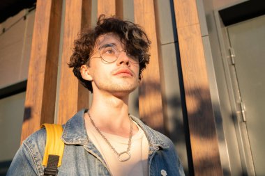 Kot ceketli ve tişörtlü sakin genç adam dış binanın yanında duruyor.