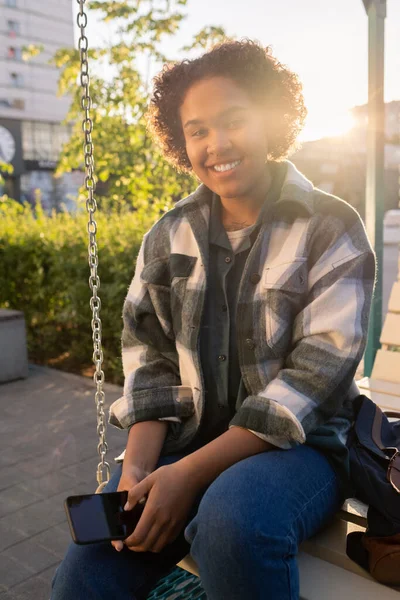 Glada tonåring med smartphone sitter på swing och tittar på dig — Stockfoto