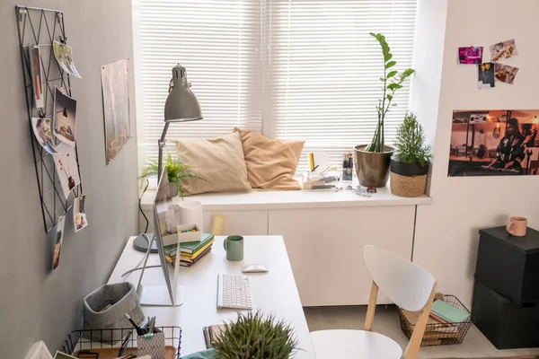 Part of room with desk by wall, chair, two cushions by window and other things — Stock Photo, Image