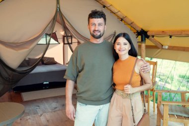 Mutlu genç adam ve kadın glamping evlerinin içinde ayakta duruyorlar.