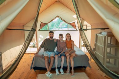 Glamping House 'da büyük bir yatakta oturan üç kişilik mutlu bir aile.