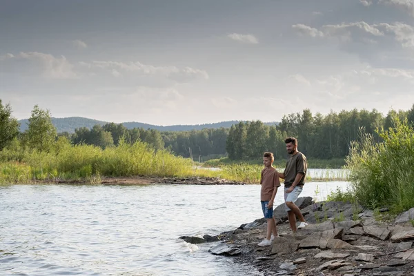 Far och son står vid sjön medan du tillbringar sommaren semester i landet — Stockfoto