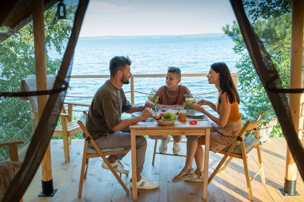 Šťastná rodina tří mají večeři u dřevěného stolu na terase — Stock fotografie