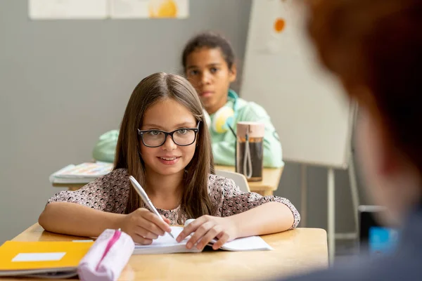 Šťastná školačka v brýlích při pohledu na učitele při psaní poznámek — Stock fotografie