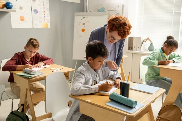 Teacher bending over desk of schoolboy during individual work — Stock Photo, Image