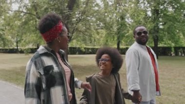 Orta halli, neşeli küçük Afrikalı Amerikalı çocuk ve ailesinin yaz günü güzel bir parkta uzun yeşil ağaçlar boyunca yürüyüp eğlendiği bir sahne.