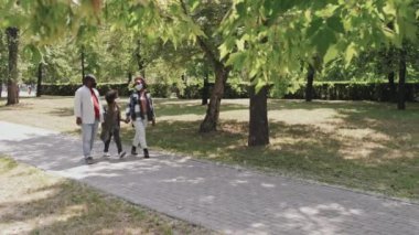 Üç çocuklu mutlu Afro-Amerikan ailesinin yavaş çekimleri parkta yürürken güneşli bir günün ve doğanın tadını çıkarıyor.