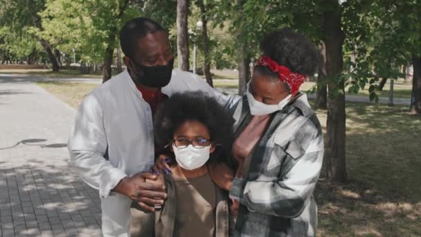 幸せなアフリカ系アメリカ人の家族の3つの着用フェイスマスクの中低速肖像夏には公園で屋外に立ってカメラのポーズ 可愛い息子を抱きしめる親 — ストック動画