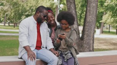 Sıcak güneşli bir günde, parkta bankta oturan ailesine akıllı telefondan fotoğraf gösteren tatlı Afro-Amerikan bir çocuğun orta yavaş çekimleri.