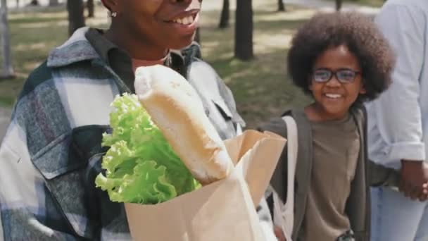 在阳光明媚的日子里 慢吞吞地用纸袋提着食品的非洲裔美国男人和女人在回家的路上和他们可爱的儿子聊着天 — 图库视频影像