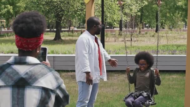 慢镜头的快乐的非裔美国人家庭发送夏天在公园外面的时间 父亲用链条摆动着他的儿子 而母亲则用智能手机给他们拍照 — 图库视频影像