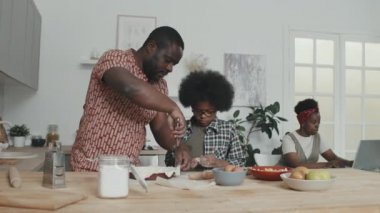 Modern Afro-Amerikan aile haftasonunu evde birlikte geçirirken çekilmiş yavaş bir fotoğraf. Anne arka planda dizüstü bilgisayarla uğraşırken baba ve oğul mutfak masasında turta yapıyorlar.