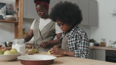 Tatlı Afrikalı Amerikalı çocuğun elmalı turta için taze elma keserken ve annesinin modern mutfakta bir arada durmasına yardım ederken ki yavaş çekimi.