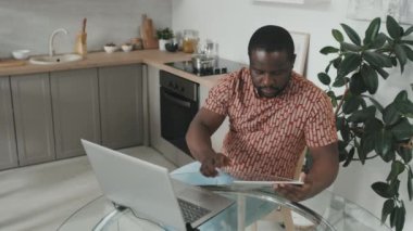 Genç Afrikalı Amerikalı adamın bilgisayarında çalışırken dijital tablet kullanırken orta yavaş çekimleri modern mutfakta masada otururken.