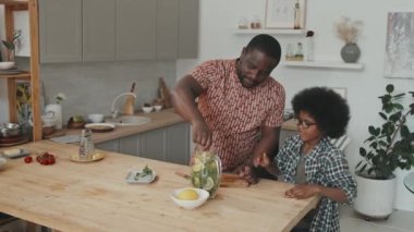 Neşeli Afrikalı Afrikalı Amerikalı baba ve oğul taze ev yapımı limonata yapıyorlar. Modern mutfaktaki cam sürahi içindeki tüm malzemeleri karıştırıyorlar.