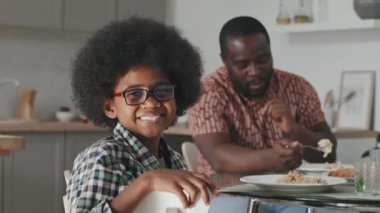 Babasıyla yemek masasında oturan ve kameraya gülümseyen Afro-Amerikalı yavaş bir çocuğun orta boy yakın portresi.