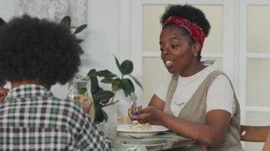 Orta yavaş çekimde, küçük oğluyla akşam yemeği yerken konuşan Afro-Amerikalı genç bir kadın.