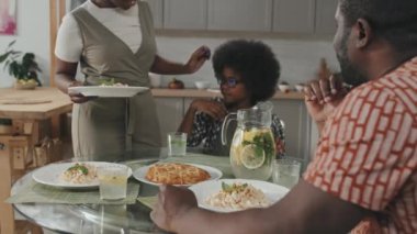 Üç kişilik modern Afro-Amerikan ailesinin yavaş çekimleri yemek masasında gözleri kapalı, el ele tutuşmuş ve yemekten önce dua ediyorlar.