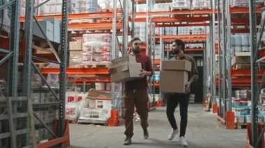 Karton kutular taşıyan neşeli erkek işçilerin yavaş yavaş izlenmesi ve rafların parçalarıyla depoda yürürken sohbet edilmesi.