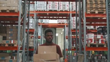 Karton kutular taşıyan Afro-Amerikalı bir adamın yavaş yavaş takibi ve rafları ürünlerle dolu bir hırdavat deposunda yürümesi.
