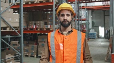 Sakallı, turuncu yelekli, kasketli, kollarını kavuşturup depoda rafları satılık eşyalarla dolu kameralara bakarken çekilmiş yavaş bir portre fotoğrafı.