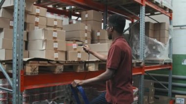 Afrika kökenli Amerikalı bir erkek depo çalışanının barkod tarayıcısı kullanarak ve raftaki karton kutuları kontrol ederek yavaş çekim görüntüleri.