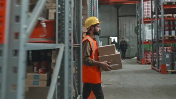 男工头戴硬帽 身穿橙色背心 背着纸板箱 走过仓库的慢镜头跟踪侧视图 — 图库视频影像