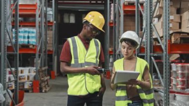 Afrika kökenli Amerikalı bir adam ve beyaz bir kadının ağır çekimde ağır çekimde takılıp kalın başlık ve yelek giymesi elinde klasör ve telsiz ile depoda çalışmayı tartışırken poz vermesi.