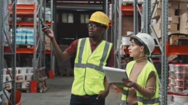 Siyah şapkalı, yansıtıcı yelek giyen, elinde telsiz olan ve elinde dosyayla beyaz kadın meslektaşıyla konuşan Afrika kökenli Amerikalı bir işçi. Depodaki raflarda bulunan eşyaları tartışıyorlar.