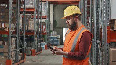 Depoda duran ve raflarda istiflenmiş eşyalara bakarken kasketli ve güvenlik yeleği giymiş sakallı erkek işçinin yavaş yavaş takibi.