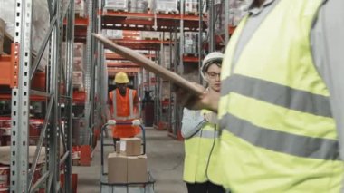 Geniş bir depoda çalışan güvenlik yelekleri ve kalın şapkalı bir grup insanın yavaş takibi. Ürünleri naklediyorlar ve envanları tartışıyorlar.