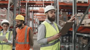 Büyük bir depoda çalışan güvenlik yelekleri ve kalın şapkalı insanların yavaş yavaş takibi. İşi, kutuları taşımayı ve stok yapmayı tartışıyorlar.