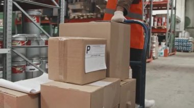 Güvenlik yelekli, yavaş yavaş tanınmayan bir erkek işçiyle orta bölüm takibi. Depoda kutuları yük arabasıyla taşıyorlar.