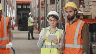 Kadın müdürün tabletli, erkek arkadaşının güvenlik yelekleri ve sert şapkalar giydiği ve kalabalık bir depoda kameraya poz verdiği yavaş çekim görüntüleri.