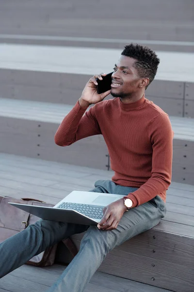 Jovem empresário contemporâneo com smartphone por orelha e laptop de joelhos — Fotografia de Stock