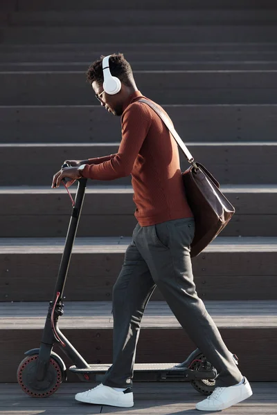 Side view of young African man listening to music in headphones while pushing scooter — Stock Photo, Image