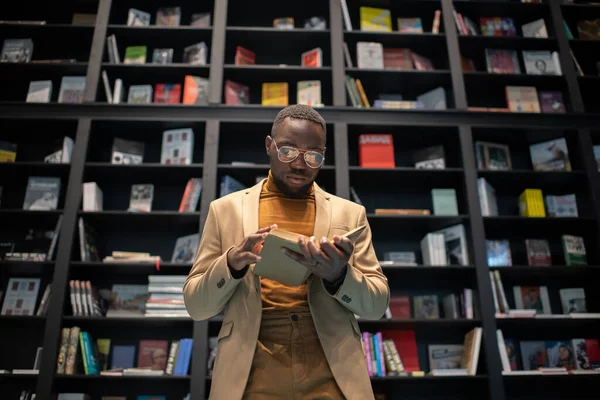 Jovem homem sério em casualwear inteligente e óculos lendo livro contra estantes — Fotografia de Stock