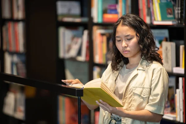 Jong serieus student lezen boek terwijl stand-by grote boekenplank — Stockfoto