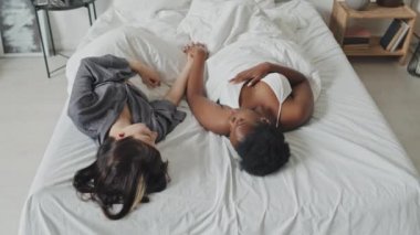 Yüksek açılı, çok ırklı lezbiyen çift yatakta uzanıp el ele tutuşurken birbirlerine sevgiyle bakıyorlar.