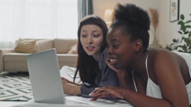 Elde tutulan, neşeli, genç, beyaz kadın ve Afro-Amerikalı kız arkadaşı rahat bir dairede uzanıp dizüstü bilgisayara bakarken sohbet ediyorlar.
