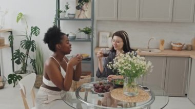 Sabahları kahve içerken rahat bir mutfakta oturup sohbet eden neşeli Afro-Amerikan kadın ve beyaz kız arkadaşının el ele tutuşması.